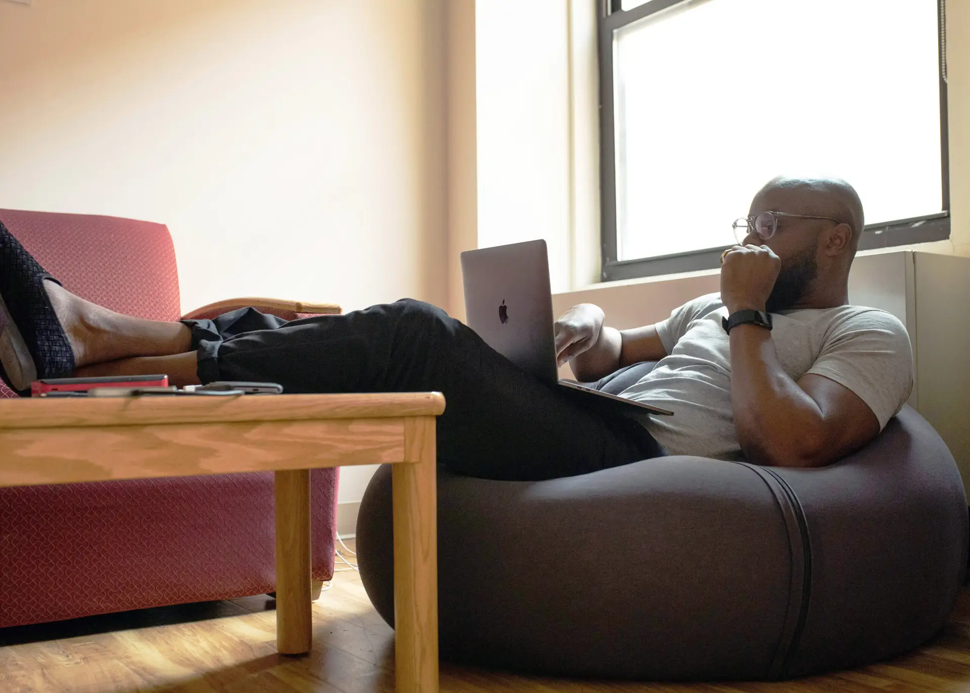 Bearded designer in glasses and wireless earbud studies a large desktop monitor, resting his chin on his hand.
