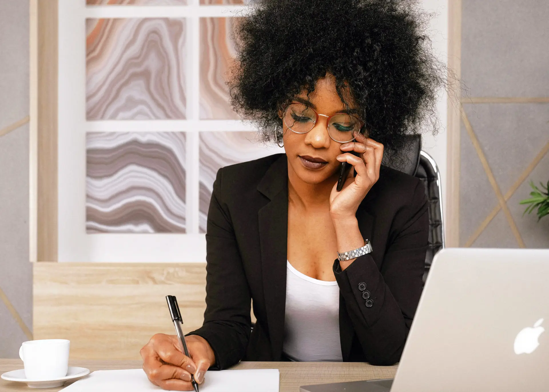 Stylish Black female consultant in blazer writes notes while phoning a client at a MacBook‑filled desk in modern studio office.