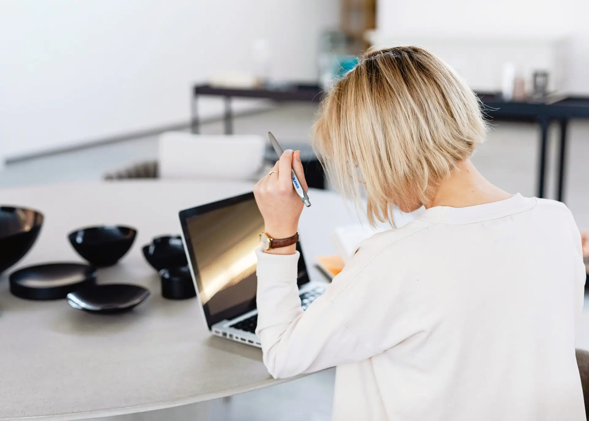 Rear view of blonde creative reviewing documents on a laptop at a minimalist studio table with matte-black ceramic prototypes.