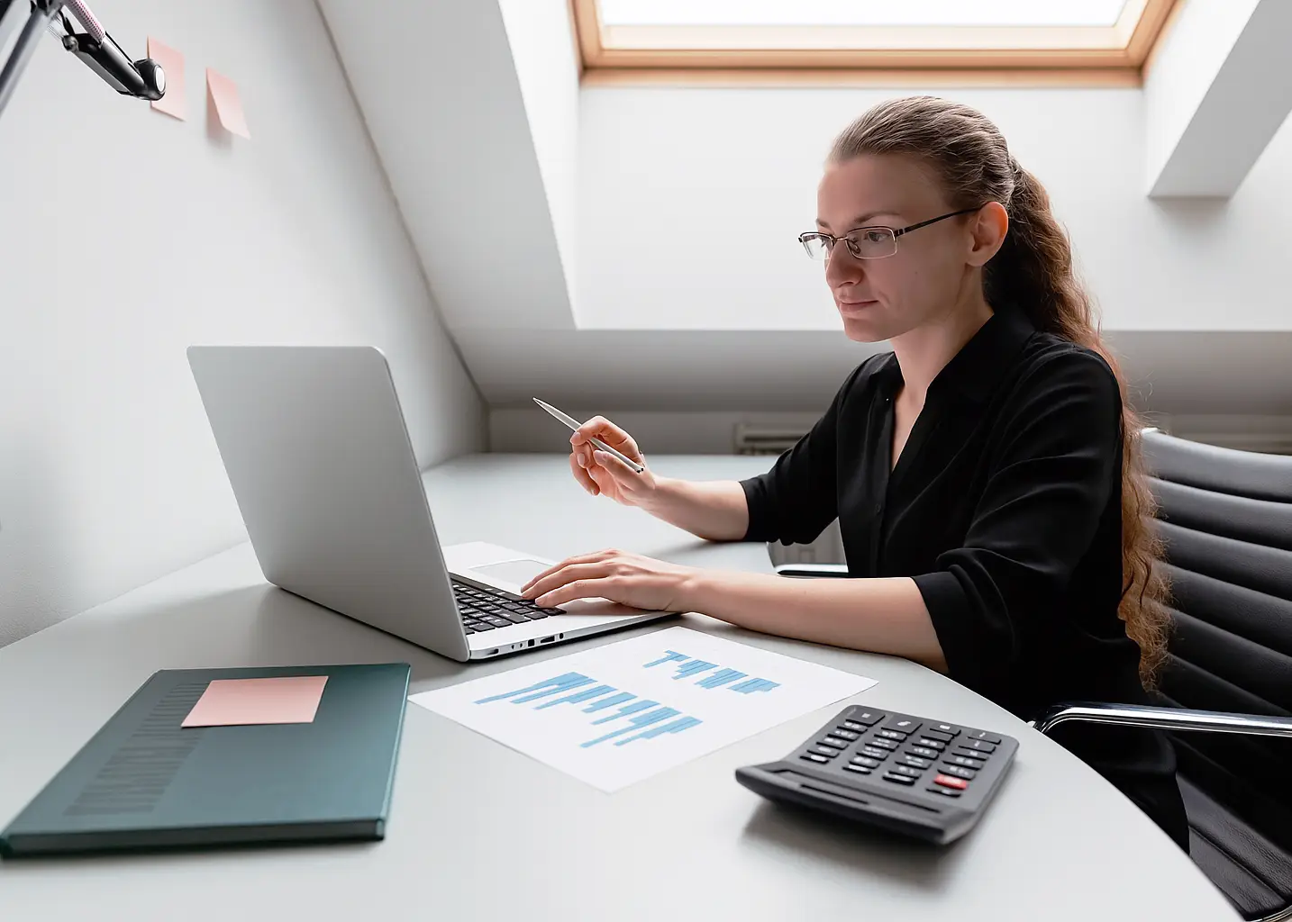 Focused female creative entrepreneur analyzes blue bar-chart printout while entering figures on a laptop in a bright loft office.