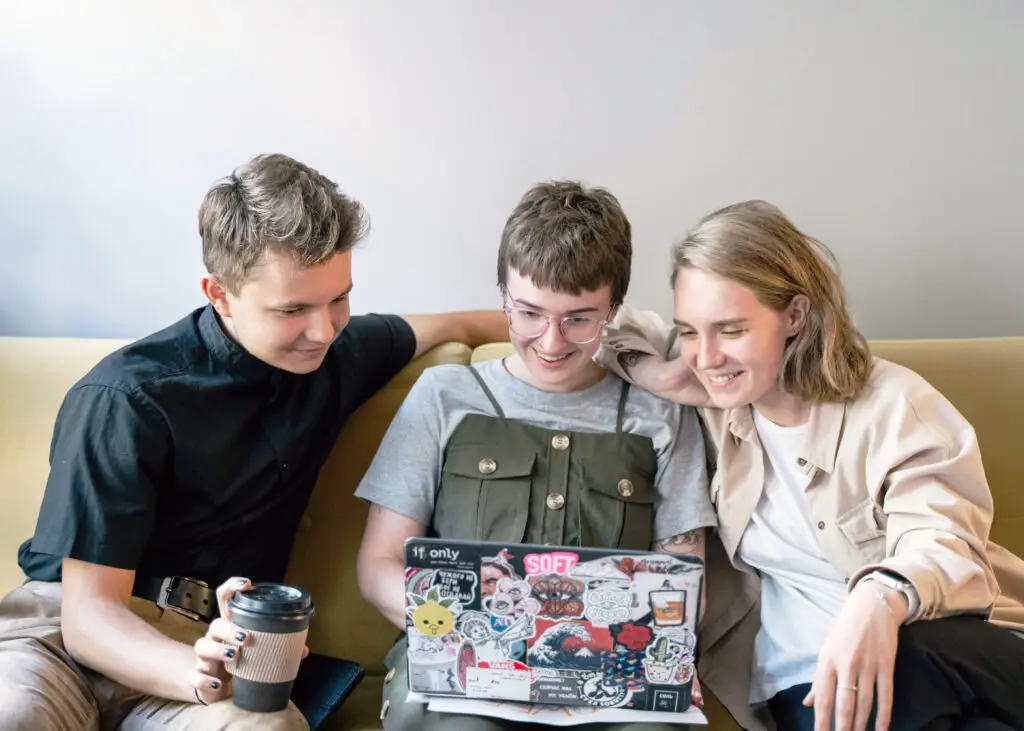 Three young creatives gathered on a sofa laugh while reviewing design-business tutorials on a sticker-covered laptop.