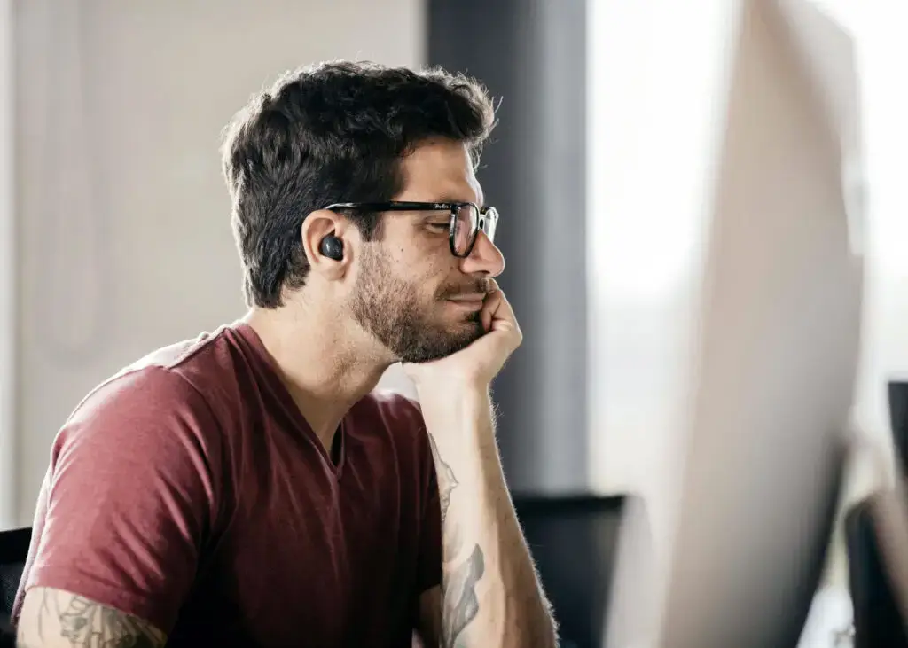 Bearded designer in glasses and wireless earbud studies a large desktop monitor, resting his chin on his hand.