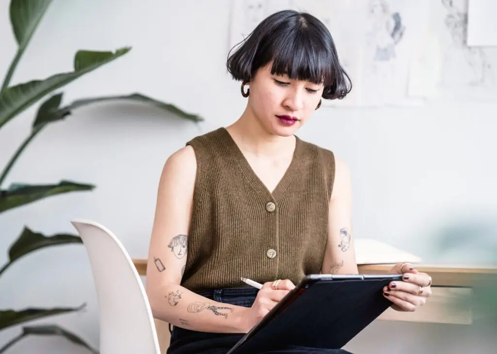Tattooed female illustrator in sleeveless knit top drafts ideas with stylus on a tablet in a minimalist studio.