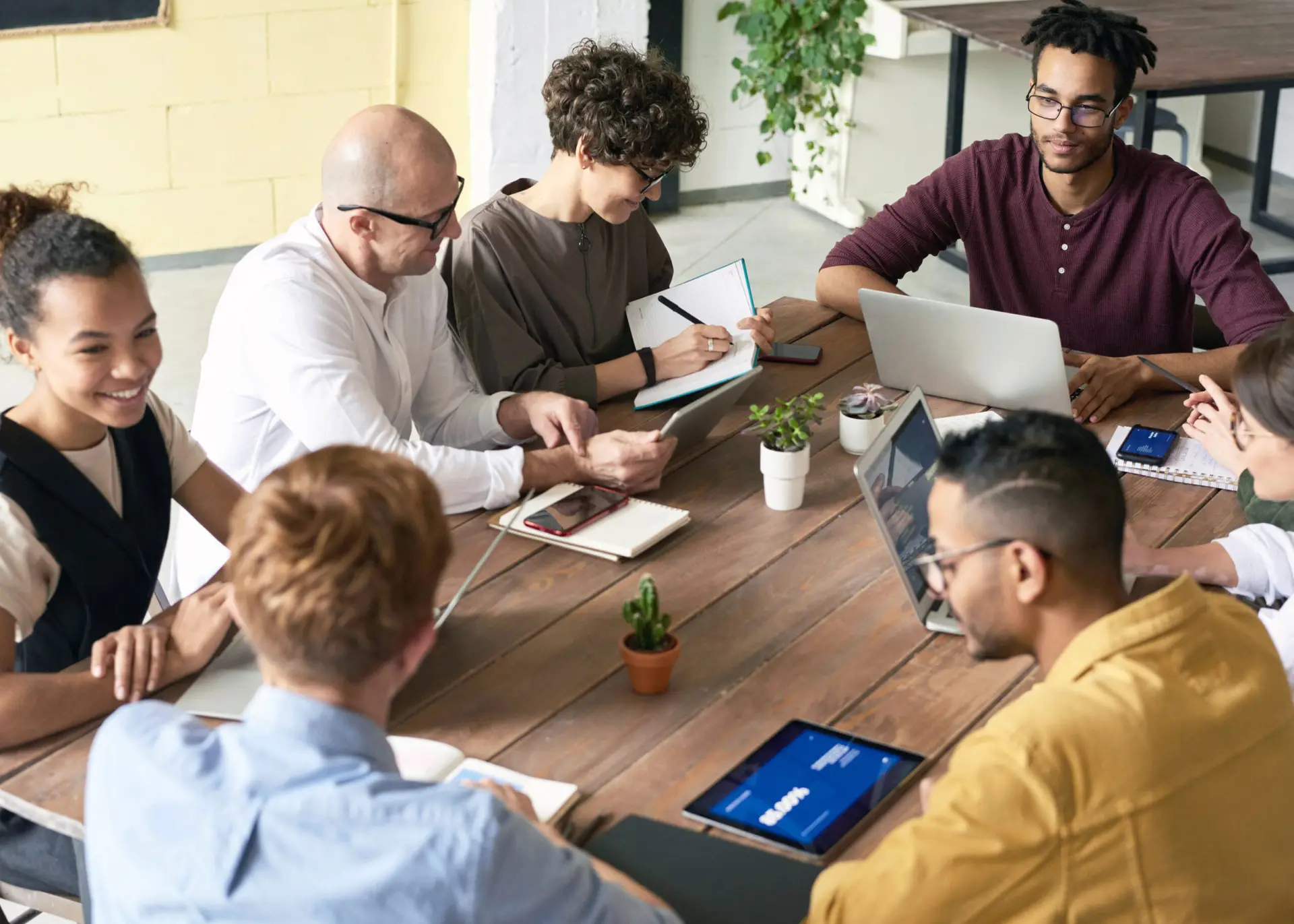 Diverse group of six creative professionals collaborating around a wooden table with laptops, tablets, and notebooks, discussing project milestones in an open-plan studio.