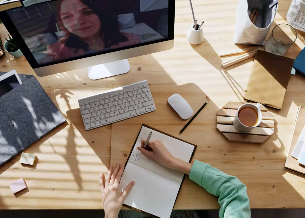 Overhead view of a home-office desk where a designer writes notes in an open notebook while chatting with a mentor on an iMac video call, surrounded by keyboard, mouse, coffee mug, and sunlight-cast shadows.