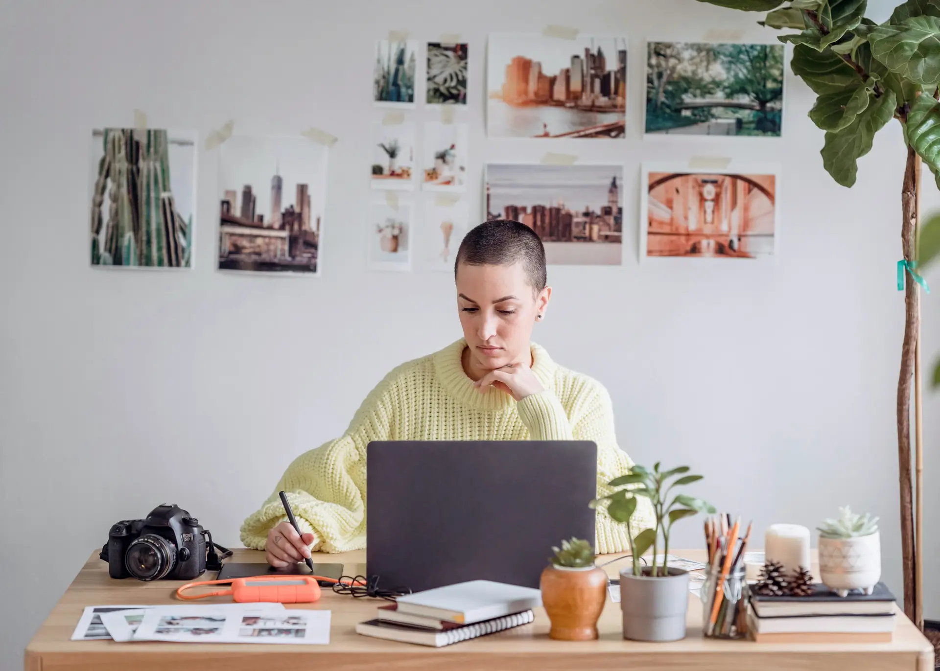 Focused designer with shaved hair in a pale-yellow sweater working on a laptop at a plant-filled desk, surrounded by sketchpads, DSLR camera, and taped inspiration photos of cityscapes on the wall.