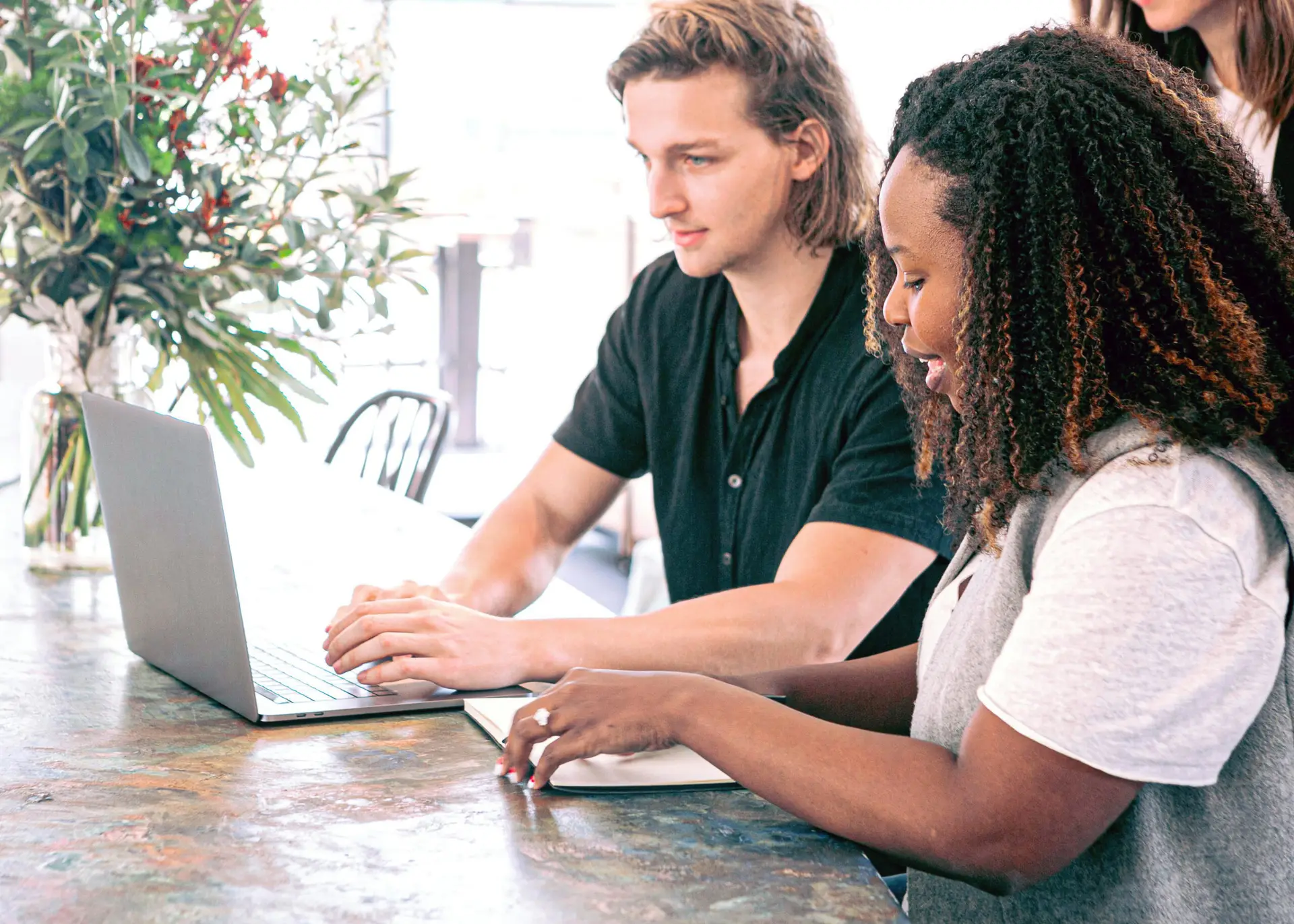 Diverse design duo brainstorms over a laptop and notebook at a sunlit coworking table with greenery in the background.
