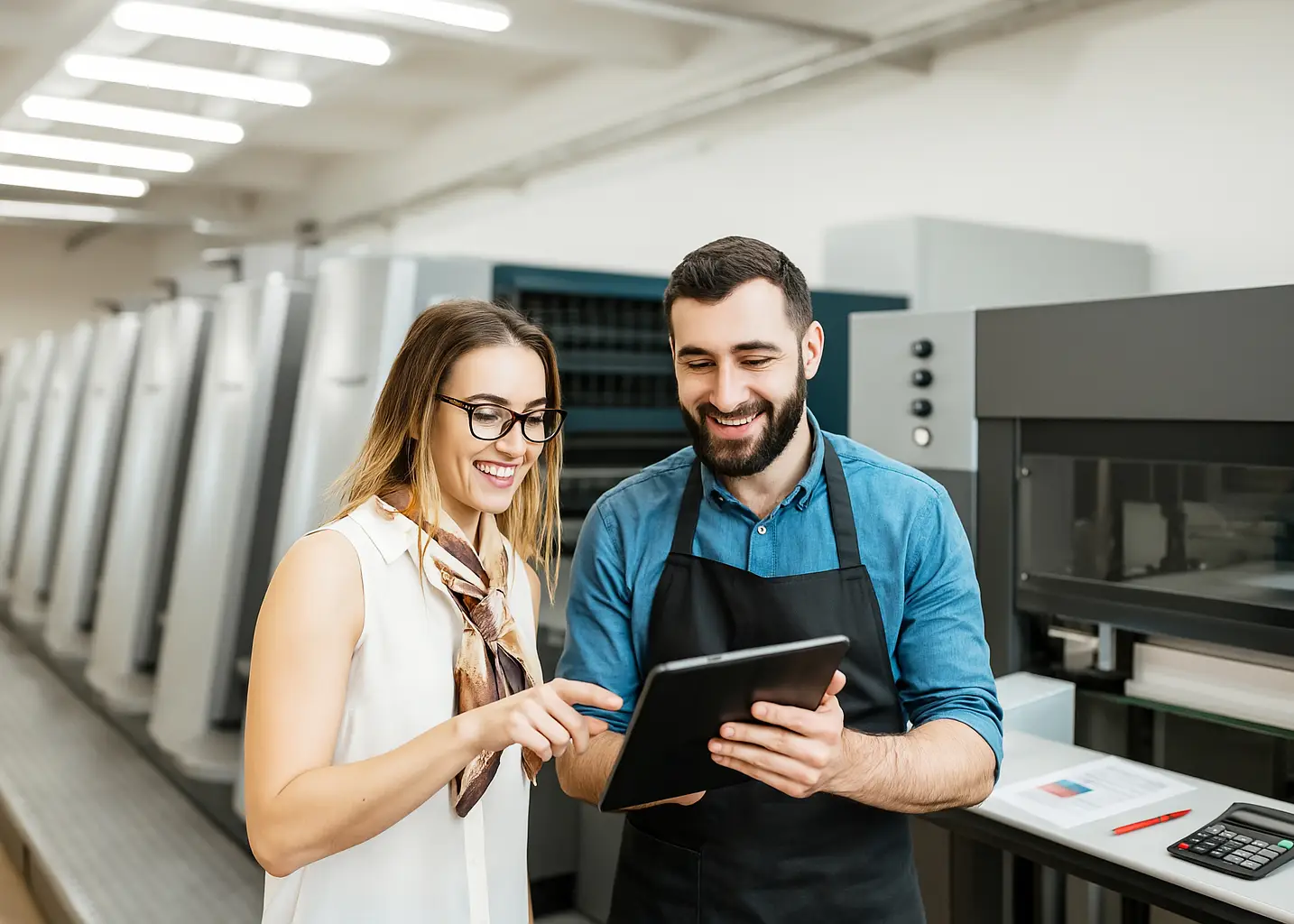 “Smiling female designer and male print technician reviewing artwork on a tablet inside a modern offset‑printing facility, surrounded by large industrial presses and production reports.”