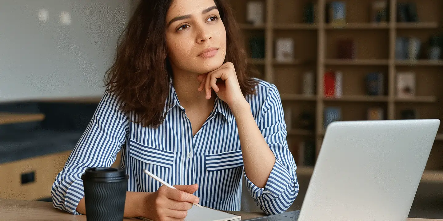Thoughtful female design entrepreneur sitting at a desk with a notebook, pen, coffee cup, and laptop, looking off-screen in contemplation.