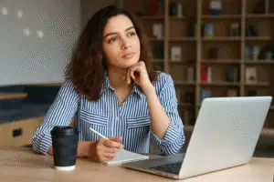 Thoughtful female design entrepreneur sitting at a desk with a notebook, pen, coffee cup, and laptop, looking off-screen in contemplation.