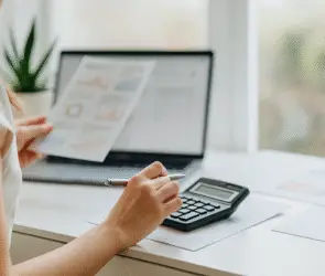 Design entrepreneur woman holding printed analytics and using a calculator at a desk with a laptop.