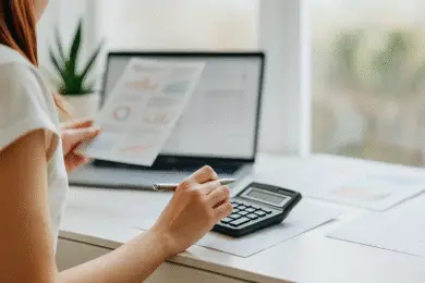 Design entrepreneur woman holding printed analytics and using a calculator at a desk with a laptop.