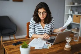 Thoughtful young design entrepreneur woman reviews financial documents and manages money using a laptop at home.
