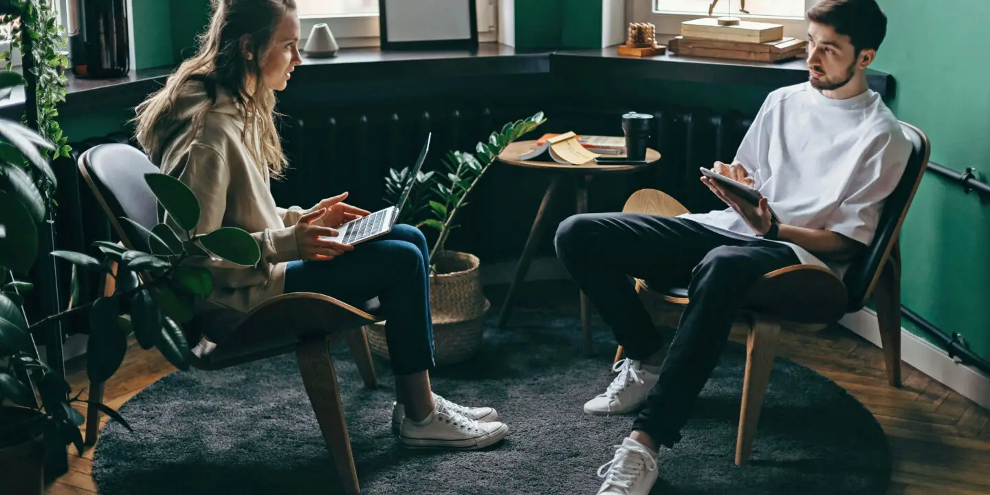 Two young design entrepreneurs discuss business ideas in a modern design studio with laptops and notepads.