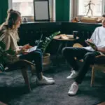 Two young design entrepreneurs discuss business ideas in a modern design studio with laptops and notepads.