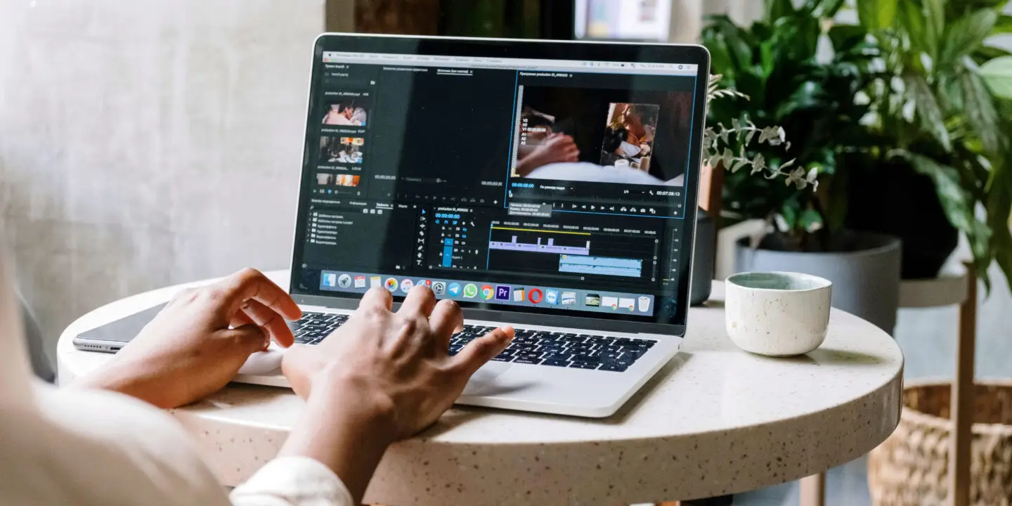 Freelance Designer editing a video project using Adobe Premiere Pro on a MacBook at a small café table with plants in the background.