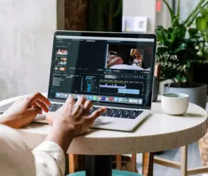 Freelance Designer editing a video project using Adobe Premiere Pro on a MacBook at a small café table with plants in the background.