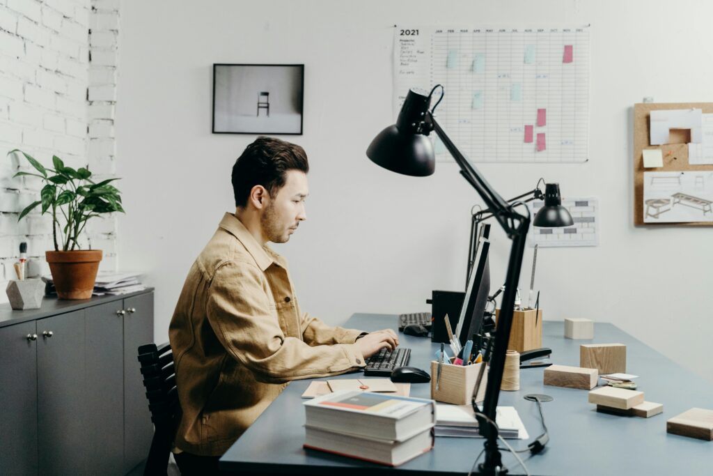 Male asian designer in a beige jacket working at a modern desk in a minimalist creative design studio with project plans and wooden prototypes.
