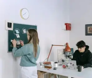 Young designer woman organizing sticky notes on a chalkboard project board while a designer man works on a laptop in a shared creative design studio.