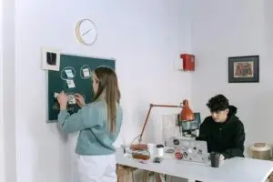 Young designer woman organizing sticky notes on a chalkboard project board while a designer man works on a laptop in a shared creative design studio.