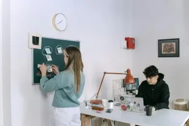 Young designer woman organizing sticky notes on a chalkboard project board while a designer man works on a laptop in a shared creative design studio.