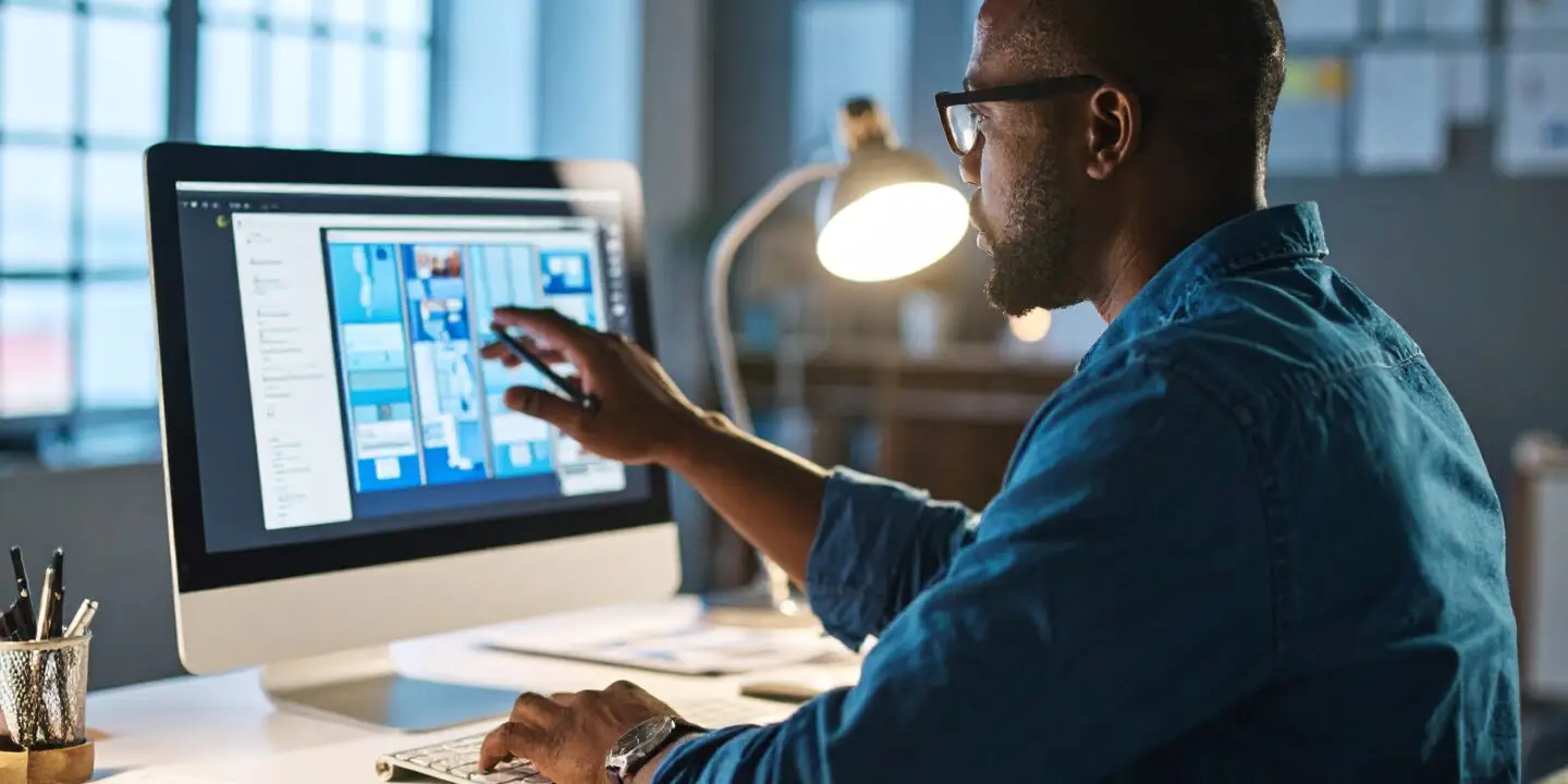 Focused male design entrepreneur creating digital wireframes for a design business website on a desktop computer in a dimly lit design studio.