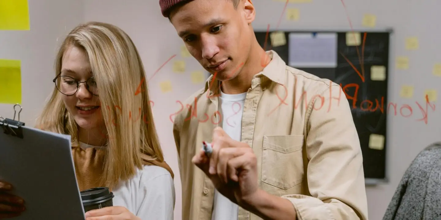 Two young design entrepreneurs brainstorming marketing strategies, writing ideas on glass with red marker while reviewing notes and holding coffee in a design studio.