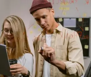 Two young design entrepreneurs brainstorming marketing strategies, writing ideas on glass with red marker while reviewing notes and holding coffee in a design studio.