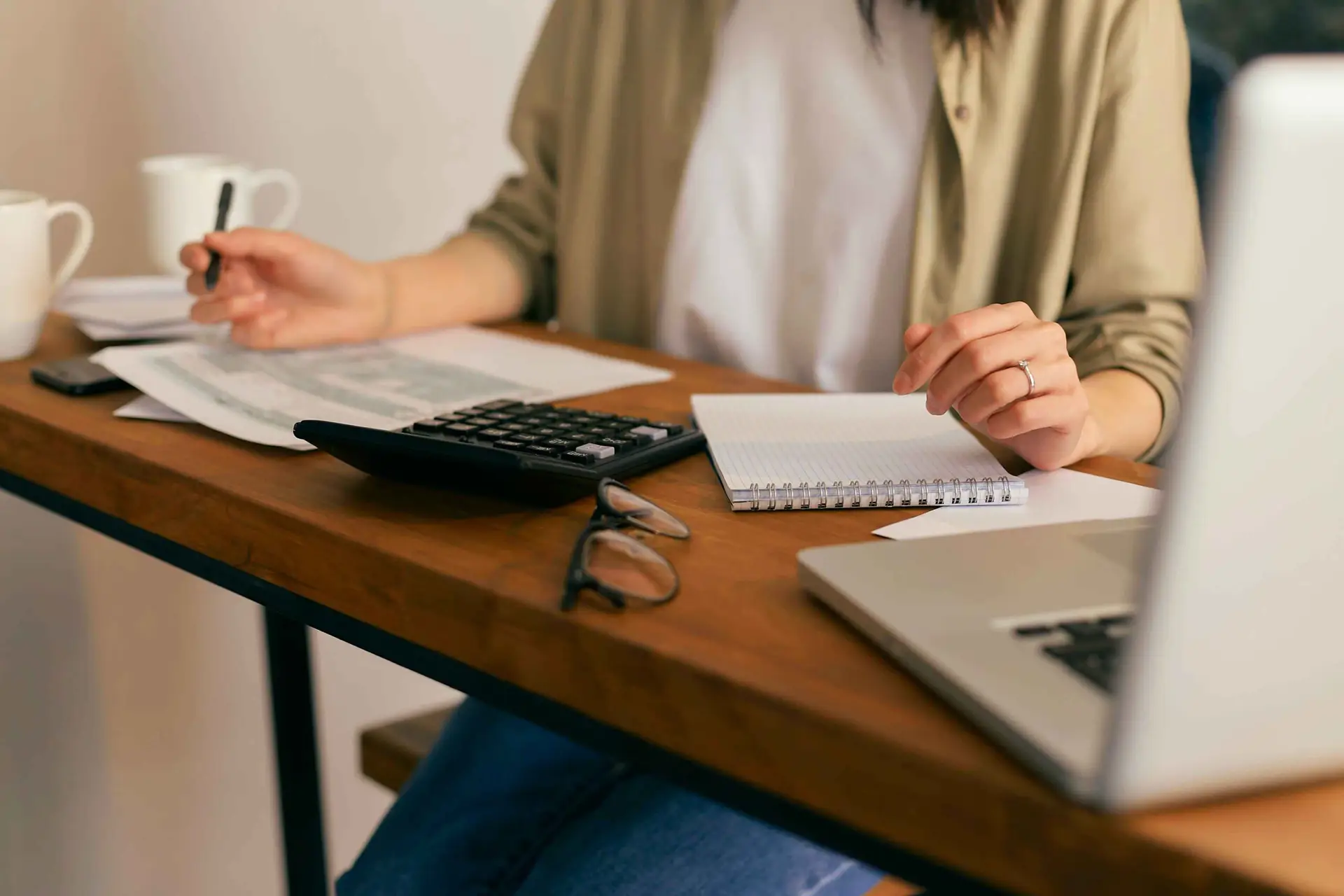 A person working at a wooden desk with a laptop, calculator, notebook, and papers, focusing on entrepreneurship and design tasks, with coffee mugs in the background.