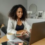 Smiling female design entrepreneur working remotely from home on a laptop with documents and coffee.