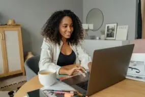 Smiling female design entrepreneur working remotely from home on a laptop with documents and coffee.