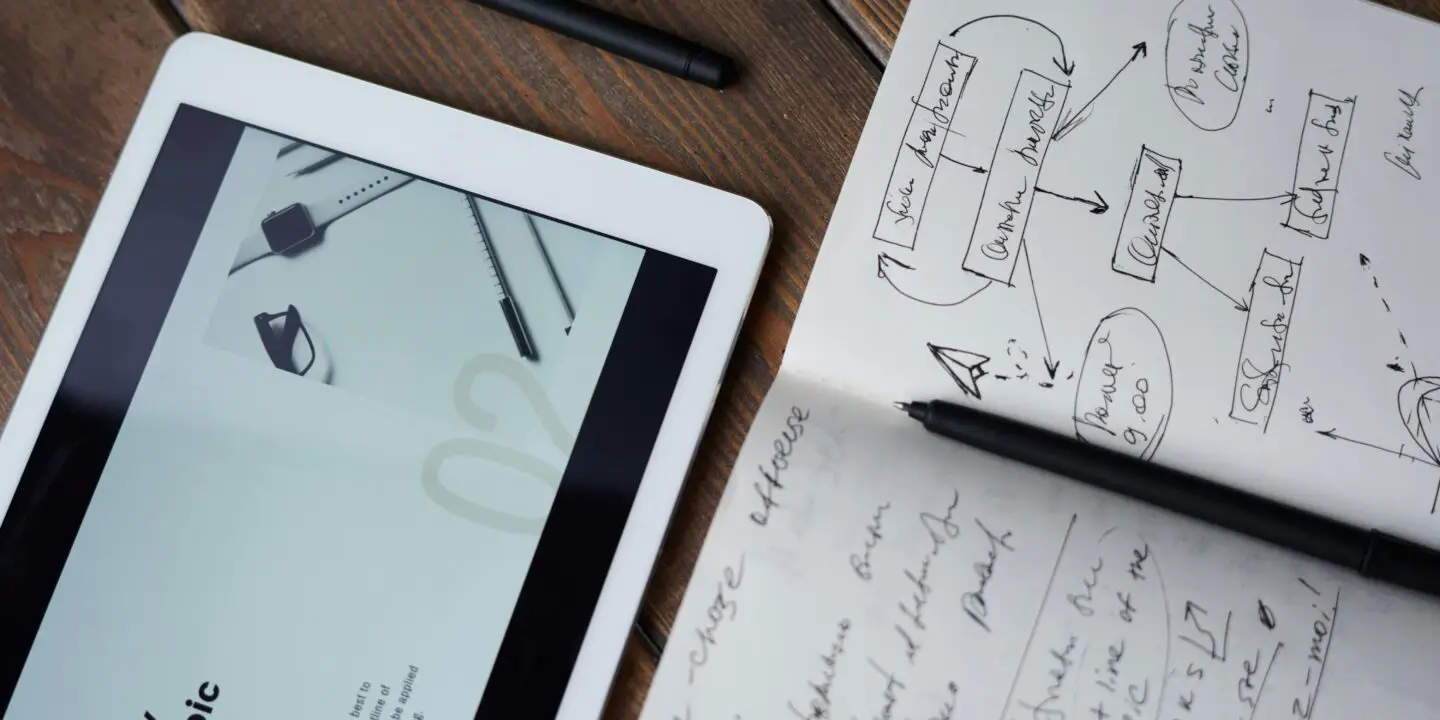 Tablet displaying design workflow content next to a sketchbook with brainstorming notes and a pen on a wooden desk