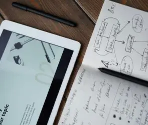 Tablet displaying design workflow content next to a sketchbook with brainstorming notes and a pen on a wooden desk