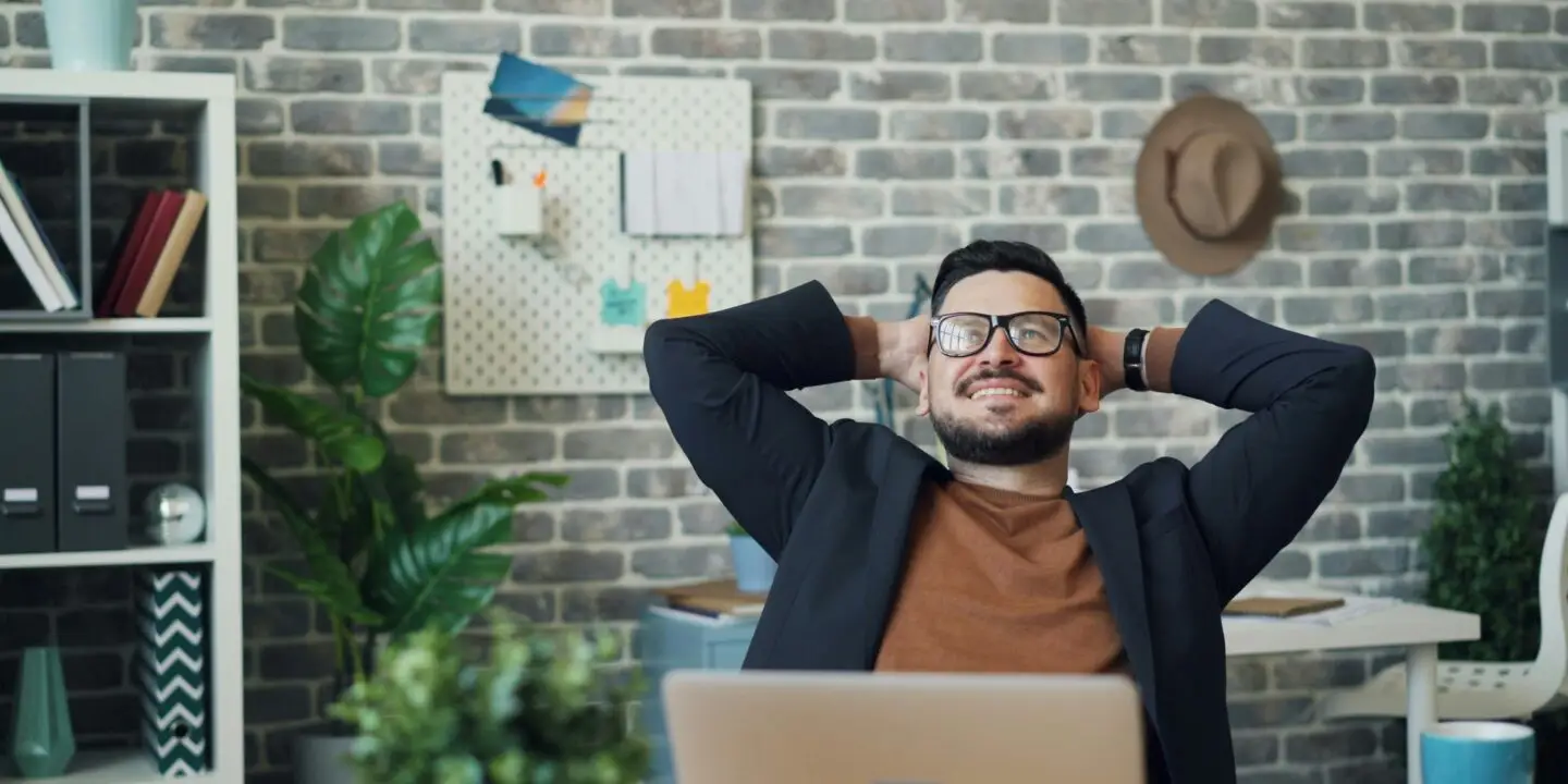 Confident male design entrepreneur relaxing in a modern office space, smiling with hands behind his head after completing a productive work session.