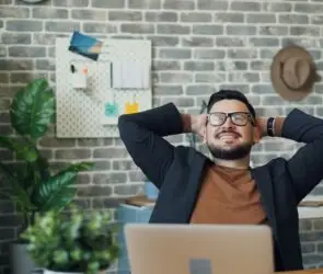 Confident male design entrepreneur relaxing in a modern office space, smiling with hands behind his head after completing a productive work session.