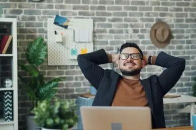 Confident male design entrepreneur relaxing in a modern office space, smiling with hands behind his head after completing a productive work session.