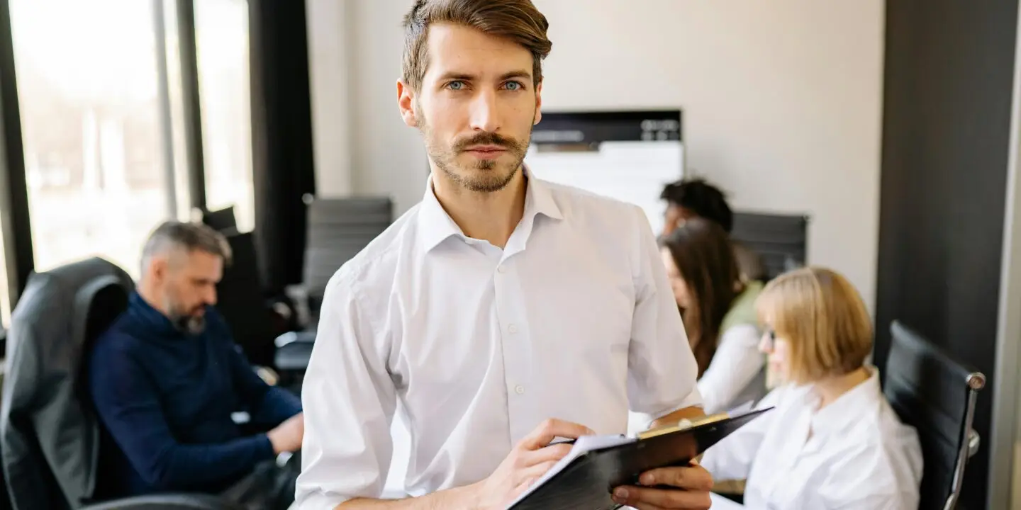 Confident male team leader holding clipboard with creative team working in the background.