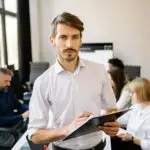Confident male team leader holding clipboard with creative team working in the background.