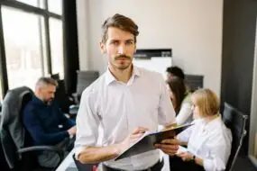 Confident male team leader holding clipboard with creative team working in the background.