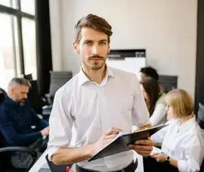 Confident male team leader holding clipboard with creative team working in the background.