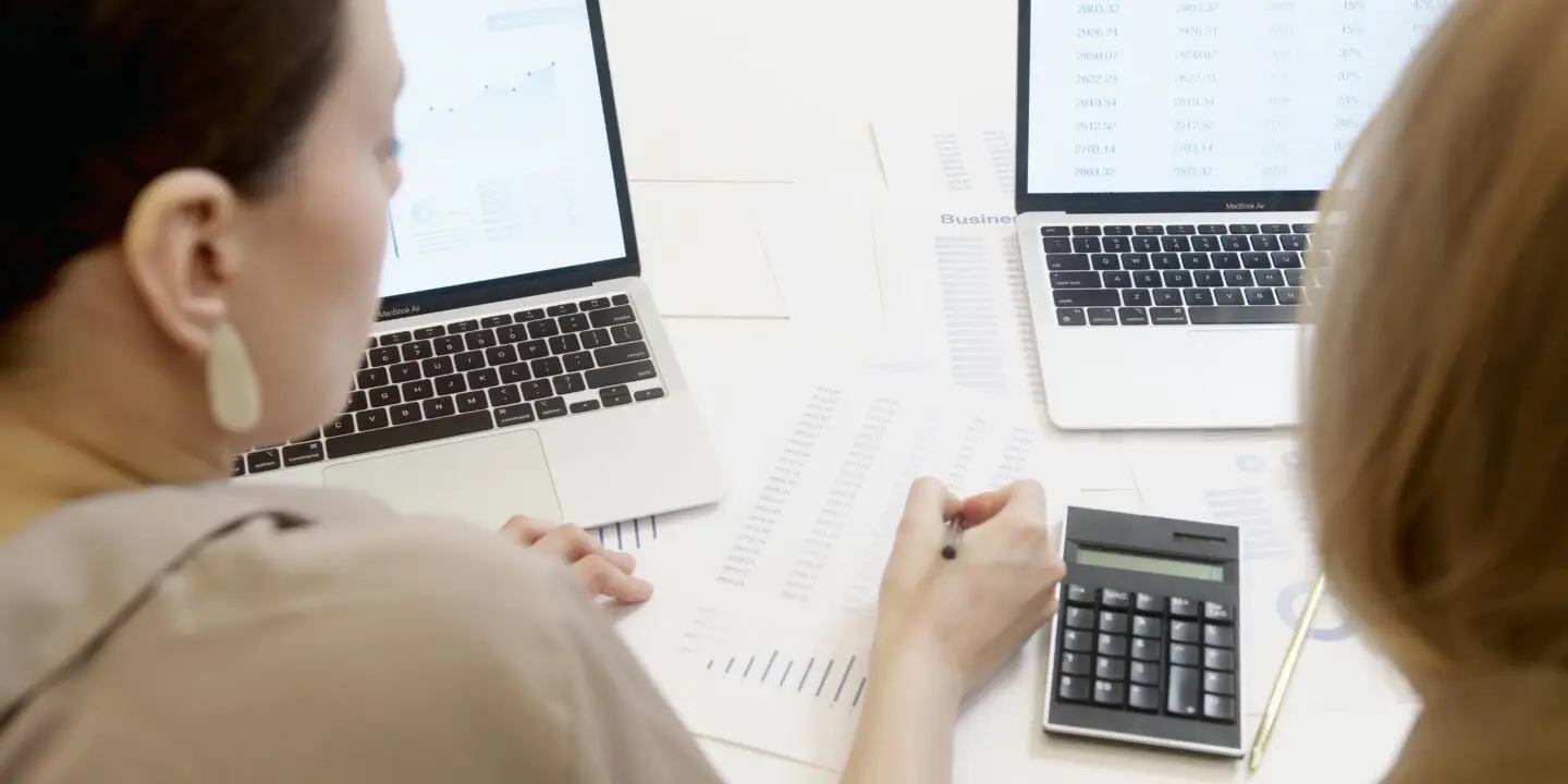 Two design entrepreneurs reviewing financial reports with laptops and calculator to manage design business budgets.