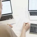 Two design entrepreneurs reviewing financial reports with laptops and calculator to manage design business budgets.