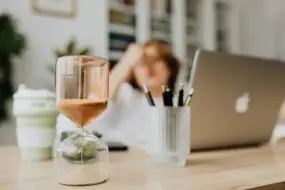 Hourglass timer on a designer’s desk with a blurred figure in the background symbolizing time management and focus for design entrepreneurs.