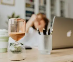 Hourglass timer on a designer’s desk with a blurred figure in the background symbolizing time management and focus for design entrepreneurs.