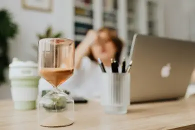 Hourglass timer on a designer’s desk with a blurred figure in the background symbolizing time management and focus for design entrepreneurs.