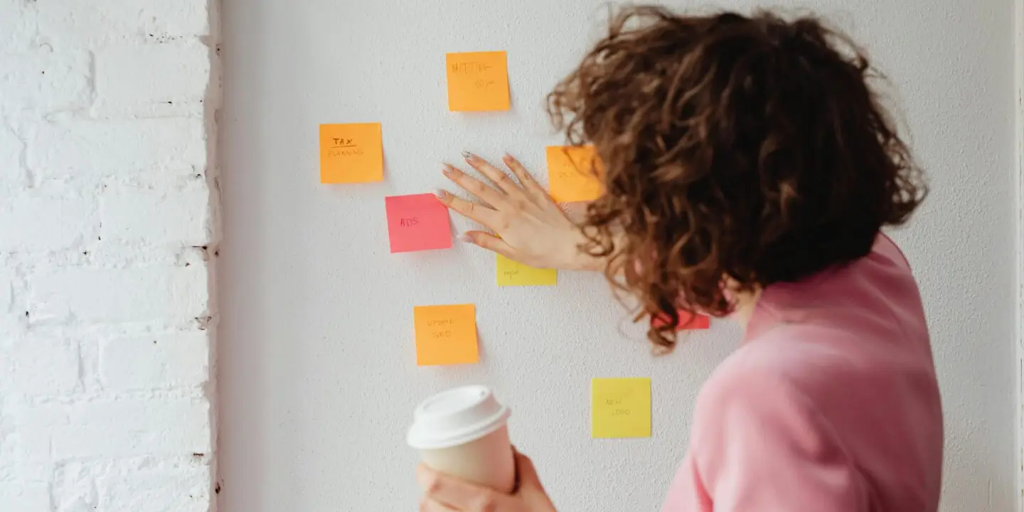 Design business woman in a pink blazer organizing colorful sticky notes on a white wall while holding a coffee cup, symbolizing design workflow planning.