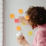 Design business woman in a pink blazer organizing colorful sticky notes on a white wall while holding a coffee cup, symbolizing design workflow planning.