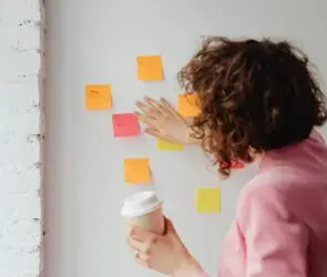 Design business woman in a pink blazer organizing colorful sticky notes on a white wall while holding a coffee cup, symbolizing design workflow planning.