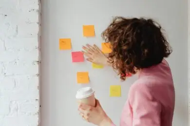 Design business woman in a pink blazer organizing colorful sticky notes on a white wall while holding a coffee cup, symbolizing design workflow planning.