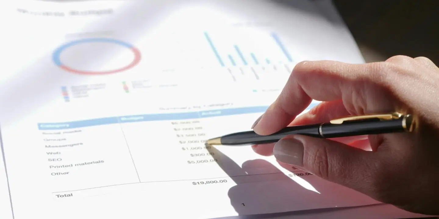 Hand holding a pen reviewing a printed design business budget sheet with graphs and charts.